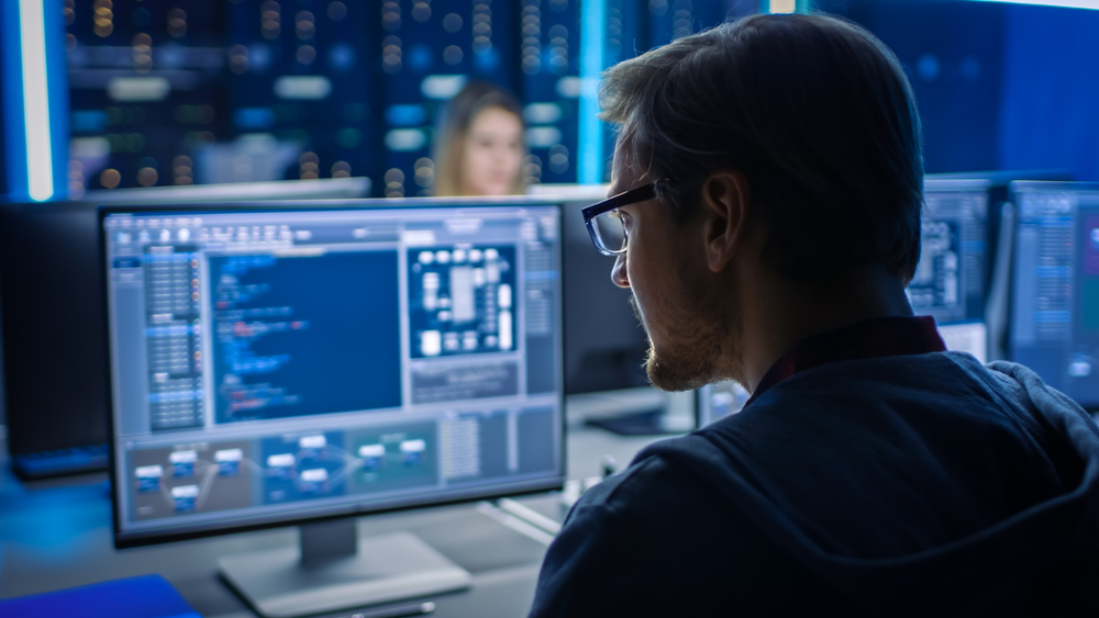 Smart Male IT Programer Working on Desktop Computer in Data Center Technical System Control Room. Team of Young Professionals Programming in Coding Language Managed IT Services in Disaster Recovery Planning